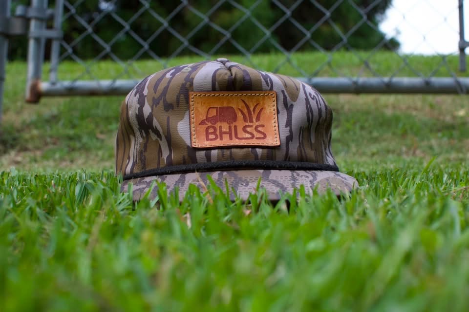 A camouflage cap with a leather BHLSS patch resting in grass in front of a chain-link fence